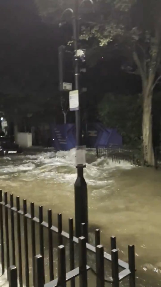 Flooded street with fast-moving, murky water filling the street past a black metal fence and lamppost.