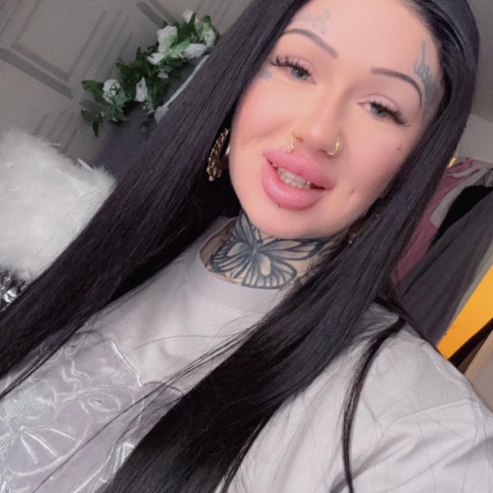 Yaz Chapman with long black hair, wearing a light-colored shirt, with tattoos on her neck and forehead, and multiple facial piercings.