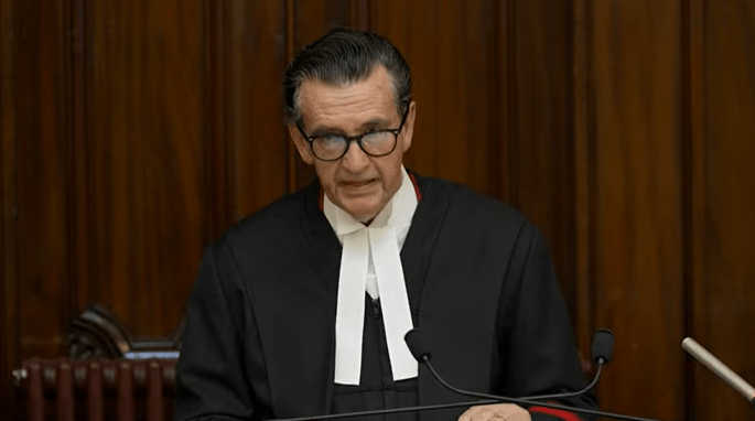 Judge in black robe and glasses speaking at a podium.