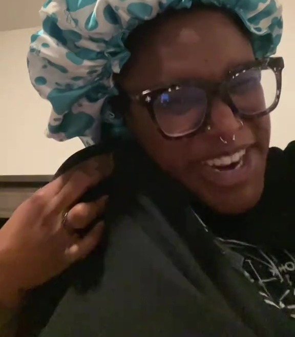 A woman in glasses and a bonnet looks at the camera, with text overlayed asking "Where are my long t!tty havers?!" and "TOUCH MY EARLOBE".