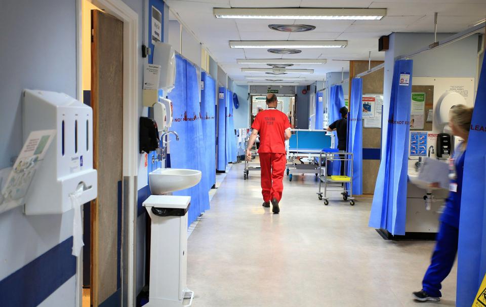 A hospital ward hallway with a medical professional in red scrubs walking away from the camera, and blue privacy curtains lining the walls.