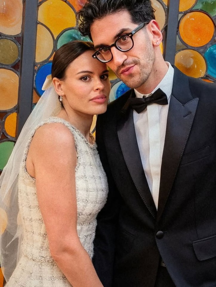 A woman in a wedding dress and a man in a tuxedo with glasses, posing together against a colorful stained-glass window.