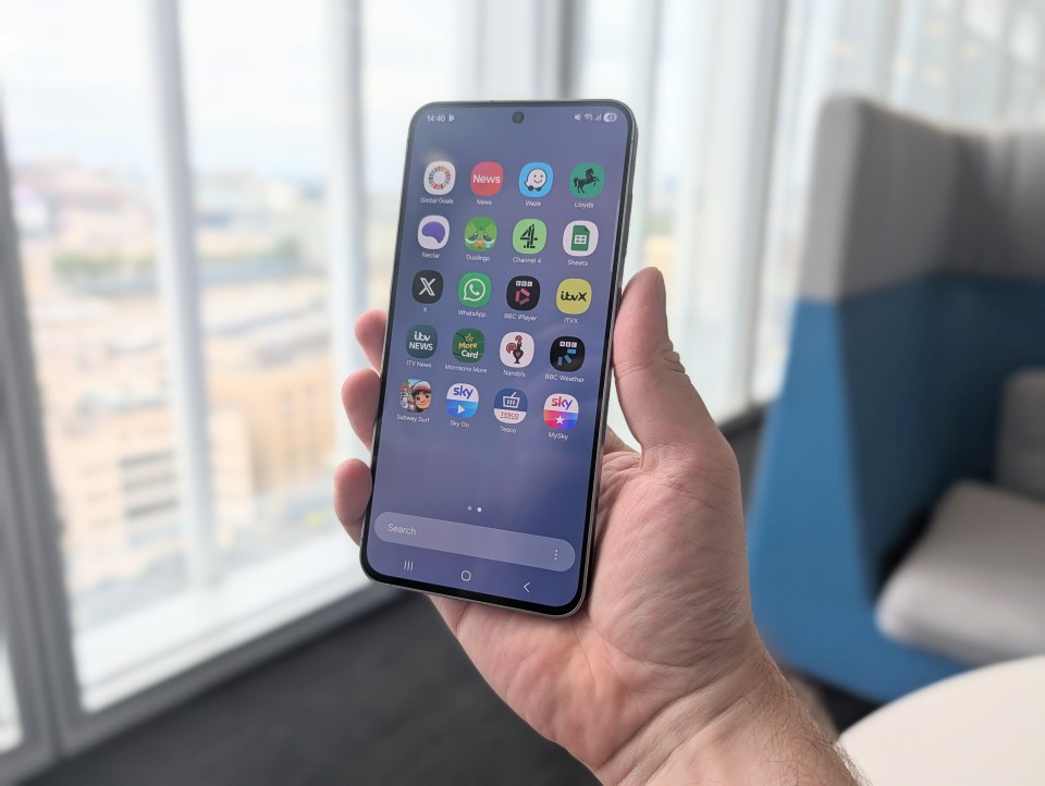 A person's hand holding a smartphone displaying various app icons including "Global Goals," "News," "Waze," "Lloyds," "Nectar," "Duolingo," "Channel 4," "Sheets," "X," "WhatsApp," "BBC iPlayer," "ITVX," "ITV News," "Morrisons More," "Nando's," "BBC Weather," "Subway Surf," "Sky Go," "Tesco," and "MySky."