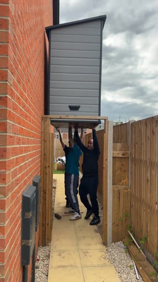 Three people struggling to move a garden shed through a narrow passage.