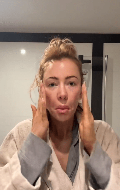 A woman with her hair in a bun applies product to her cheeks with both hands.