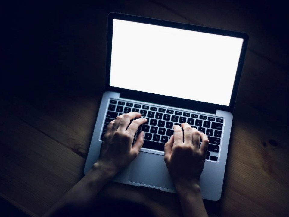 A person typing on a laptop with a blank, bright screen in a dimly lit room.