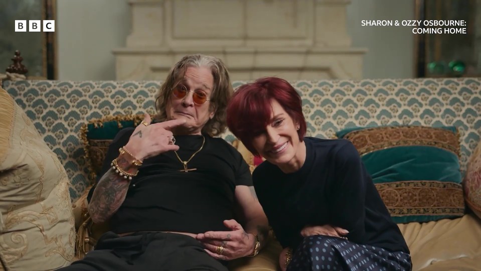 Sharon and Ozzy Osbourne sitting on a patterned couch, with Ozzy pointing at himself and Sharon smiling.