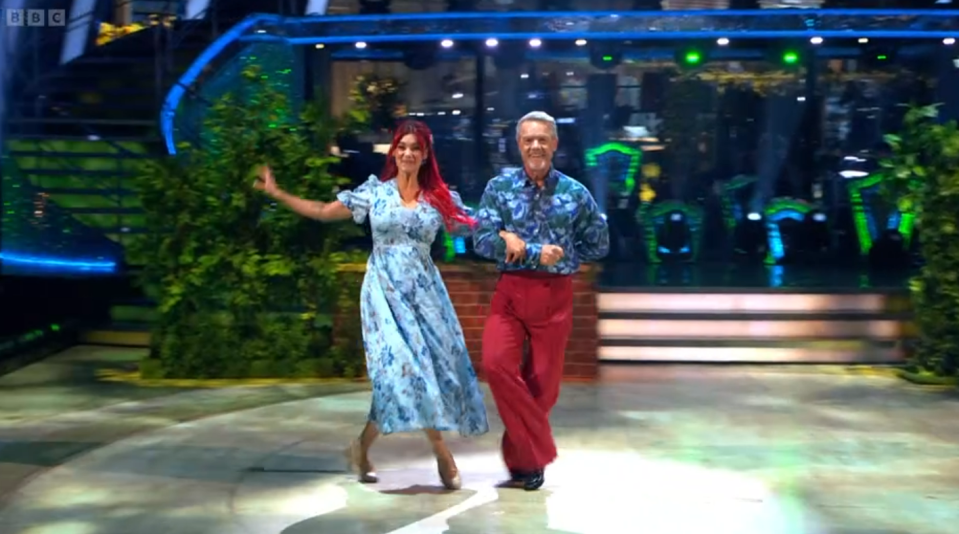 A woman in a blue floral dress and a man in a blue floral shirt and red pants dancing on a stage.