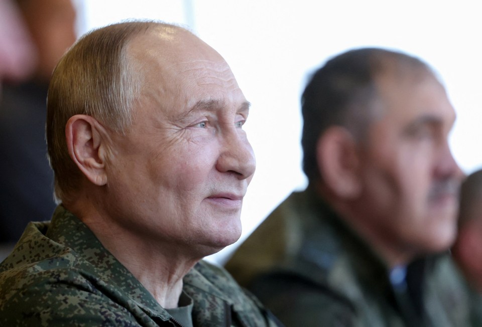 Russian President Vladimir Putin in a military uniform, looking towards the right, with another man blurred in the background.
