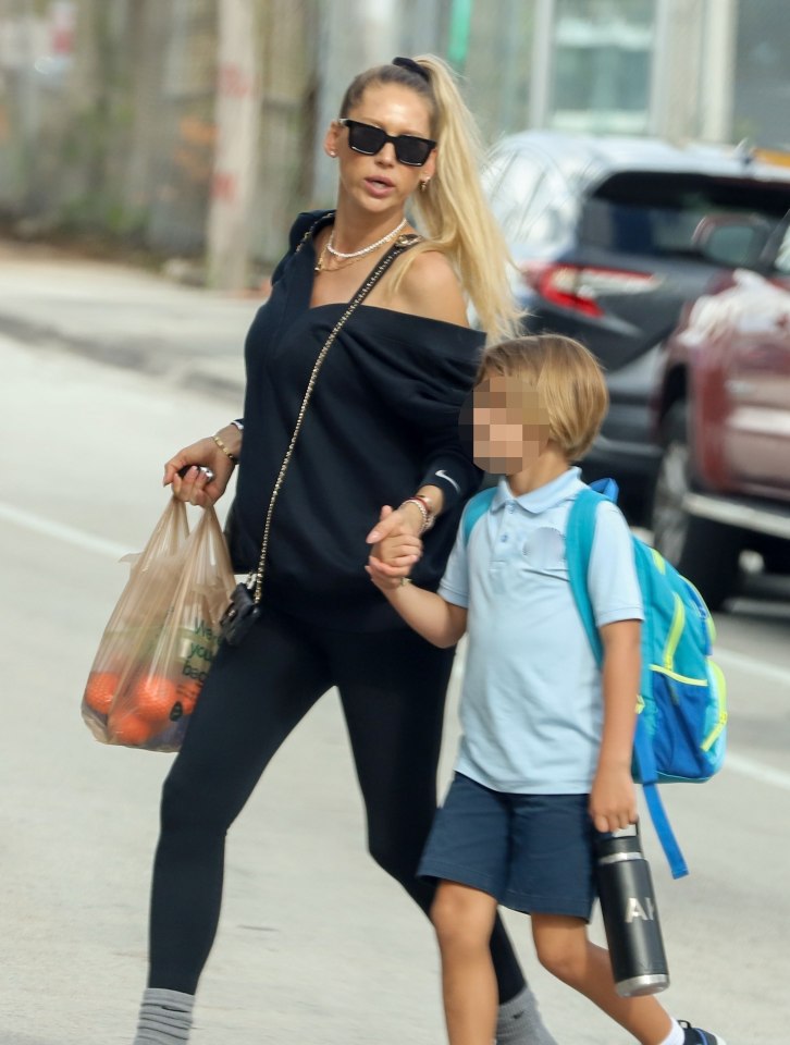 A woman and a young child walking together, the woman holding a bag of oranges and the child holding a water bottle and wearing a backpack.