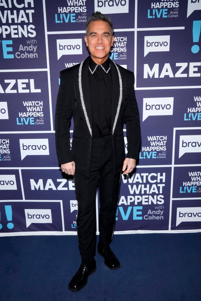 Robbie Williams smiling, wearing a black suit with sparkling silver lapels, standing in front of a blue background with "Watch What Happens Live" and "Bravo" logos.