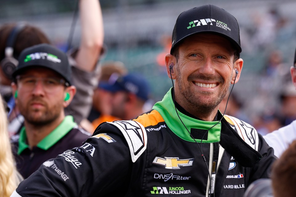 Romain Grosjean in a black and green racing suit and black Juncos Hollinger Racing hat, smiling on pit lane at Indianapolis 500 qualifying.