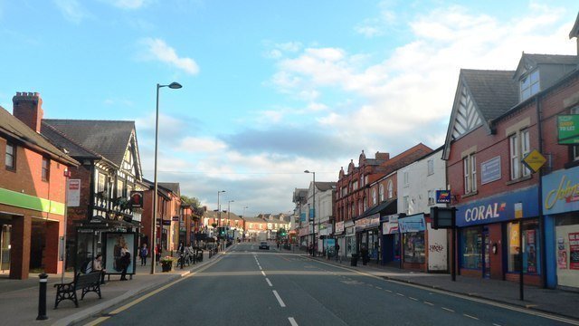 Street lined with buildings, including shops and pubs, under a partly cloudy sky.