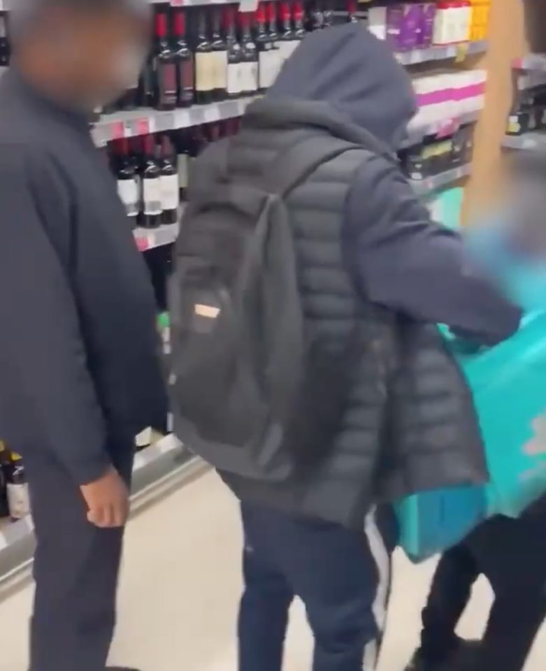 A person in a black puffer jacket and a person in a black long-sleeved shirt shoplifting bottles of alcohol into a teal bag, with a security guard standing nearby.