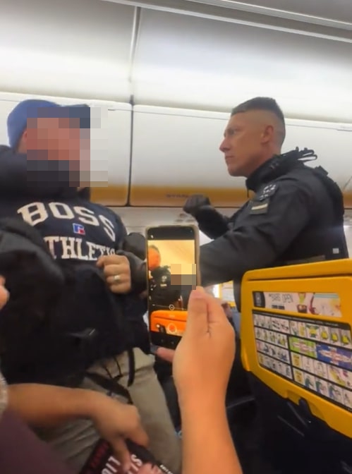 A man in a blue hat and "BOSS ATHLETIC" sweatshirt being escorted from an airplane by a police officer while another passenger records it on a phone.
