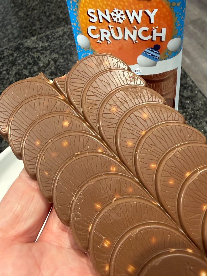 A hand holding a chocolate bar with a fan-shaped design, next to a bag of Snowy Crunch.