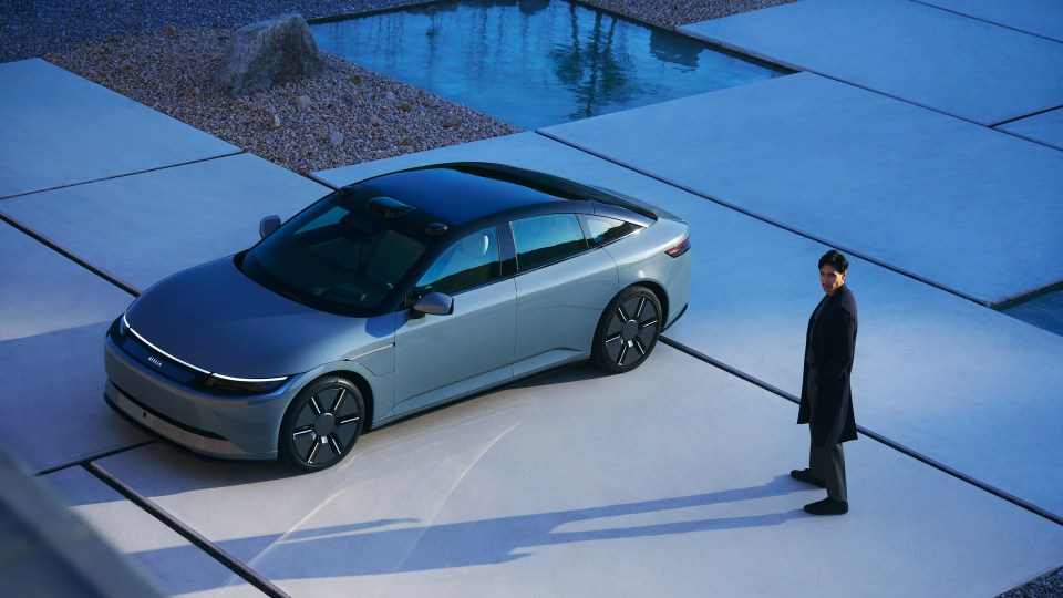 A man in a dark coat and trousers standing next to a gray Afeela car.