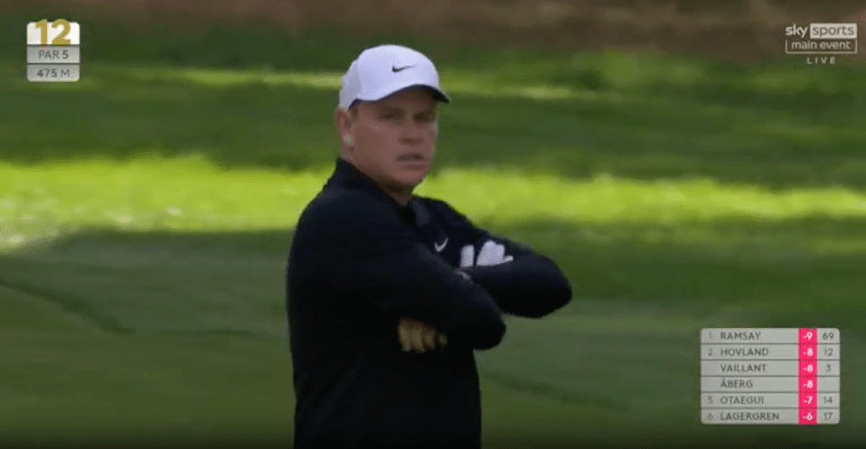 Golfer with arms crossed, scoreboard in view.