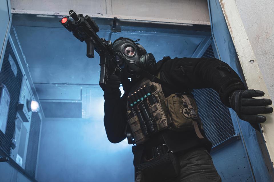 Special unit soldier with a gas mask emerges from an industrial elevator, aiming a rifle with a red laser sight.