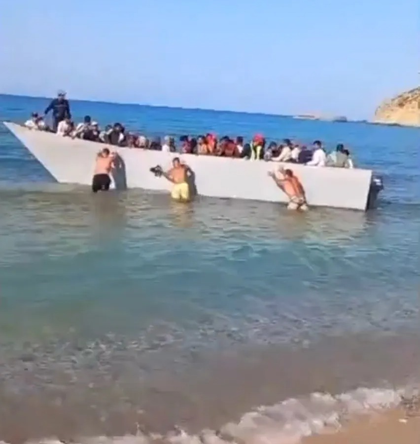 A crowded boat filled with people, with some individuals in the water, is seen near a beach, under a blue sky with Greek text overlay.