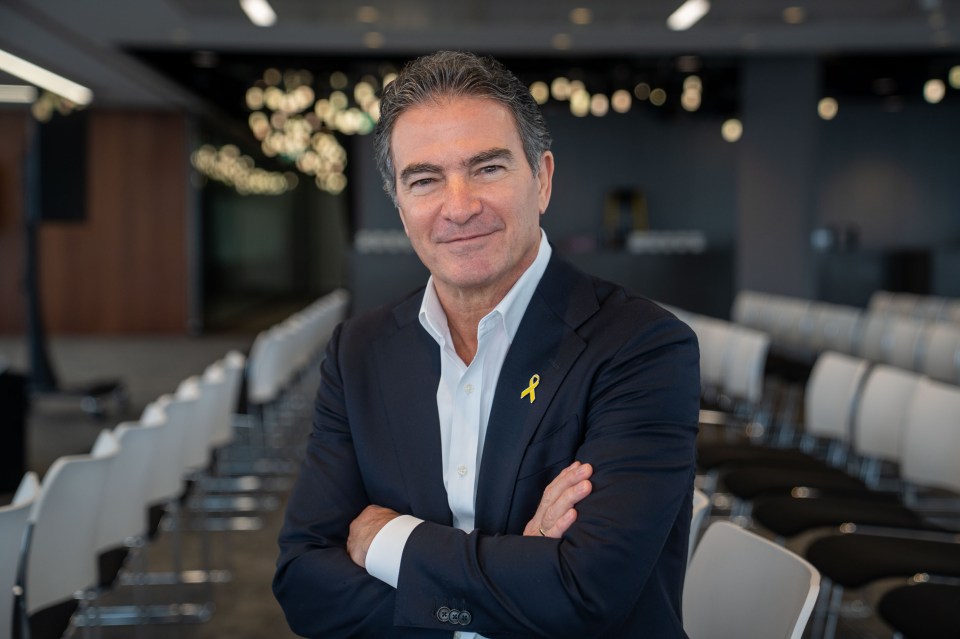 Yossi Cohen, former head of Mossad, smiling with arms crossed, wearing a suit with a yellow ribbon pin.