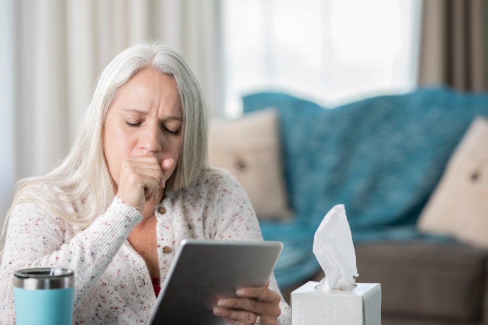 a woman coughs while using a tablet next to a box of tissues
