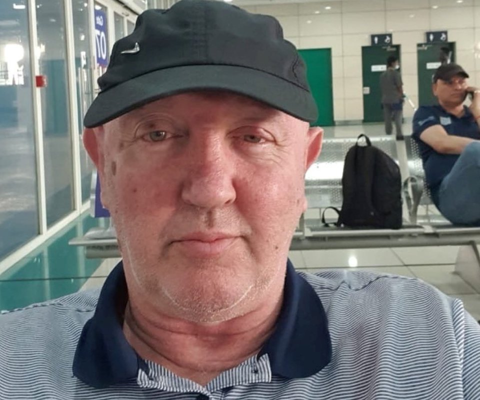 John Murphy, a British veteran, wearing a black cap and striped shirt, sitting in what appears to be an airport or public waiting area.