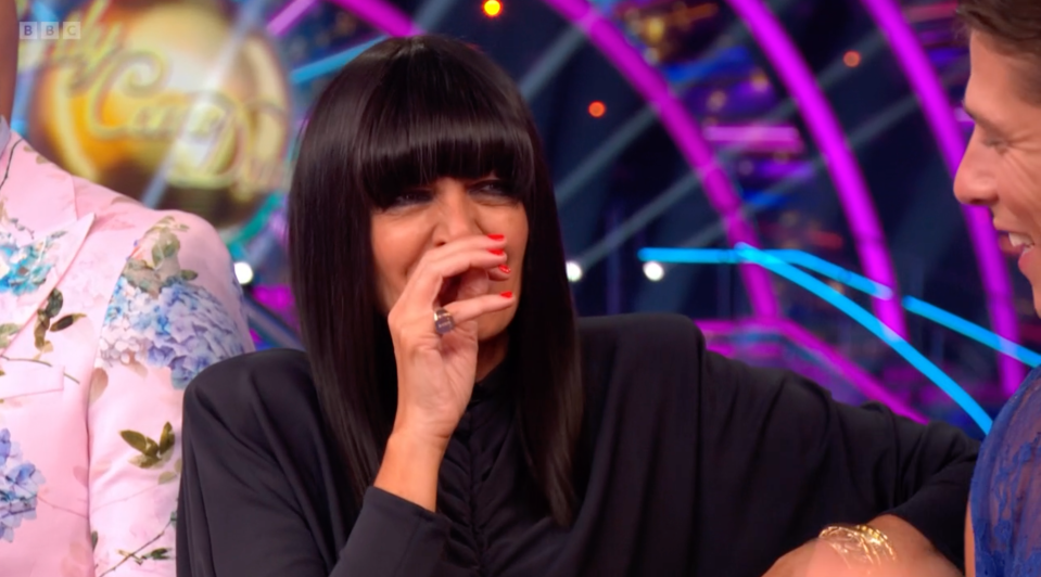 Claudia Winkleman covering her mouth and laughing while wearing a black shirt, next to a man in a floral pink jacket.