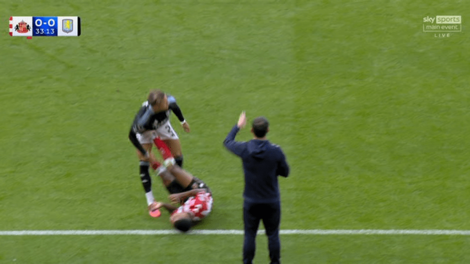 A football player in a black and white uniform appears to have tackled an opponent in a red and white uniform, who is lying on the ground. Another man in a navy blue tracksuit gestures with his hand.