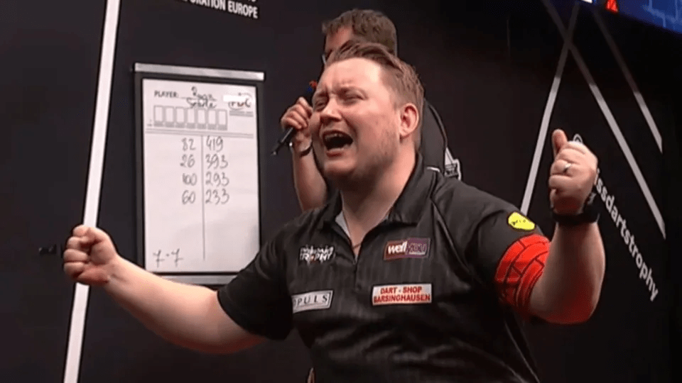 A male darts player in a black shirt celebrates a victory with his fists in the air.