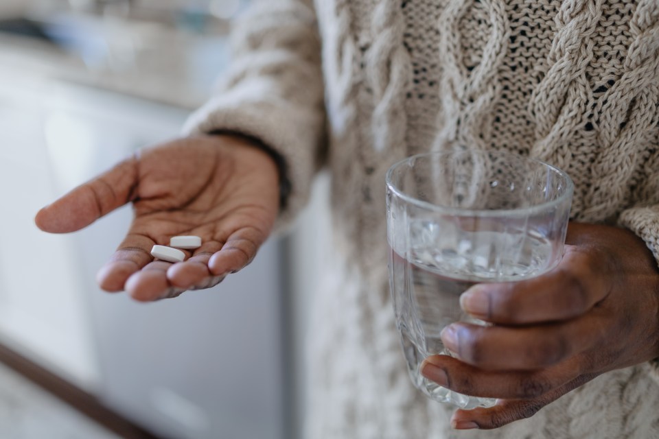 Close-up of a Black woman holding pills in one hand and a glass of water in the other.