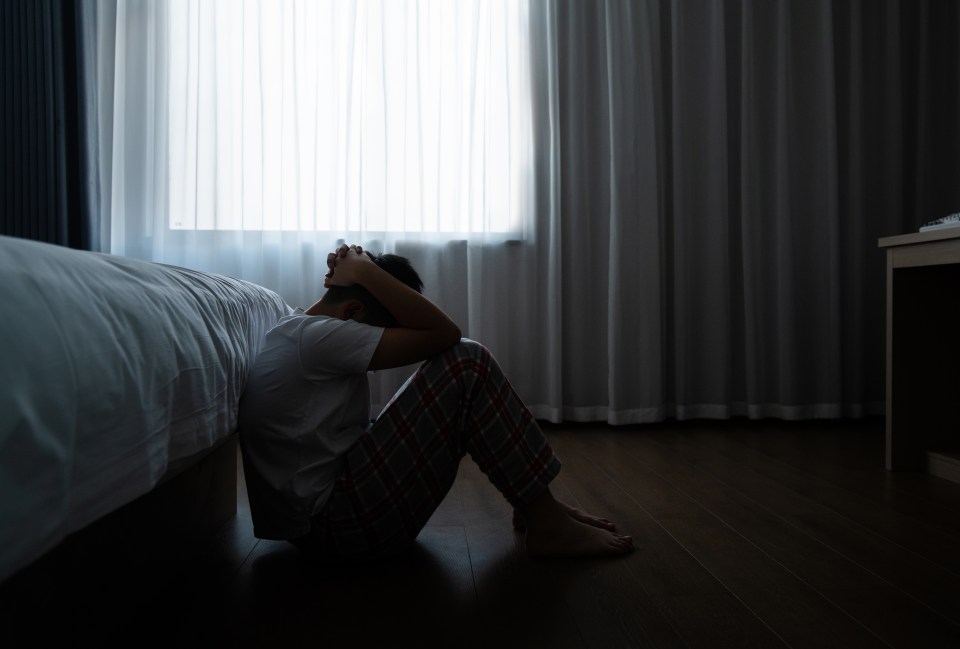 A depressed teenage boy sits on his bedroom floor, head in hands.