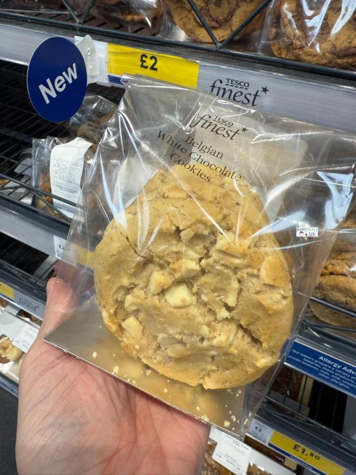 A hand holds a single Tesco Finest Belgian White Chocolate Cookie in its packaging, with a "New" sign and £2 price visible in the background.