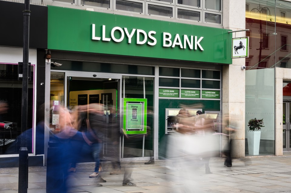 Exterior view of a Lloyds Bank branch in London with blurred people walking past, highlighting the bank's services and security warnings.