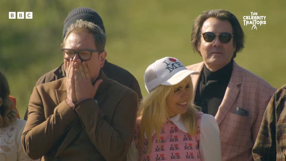 A group of people, with one man covering his mouth and a woman in a "I Love Me" hat smiling, from The Celebrity Traitors.