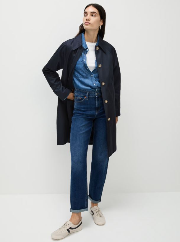 Woman wearing a navy trench coat, denim shirt, jeans, and sneakers.