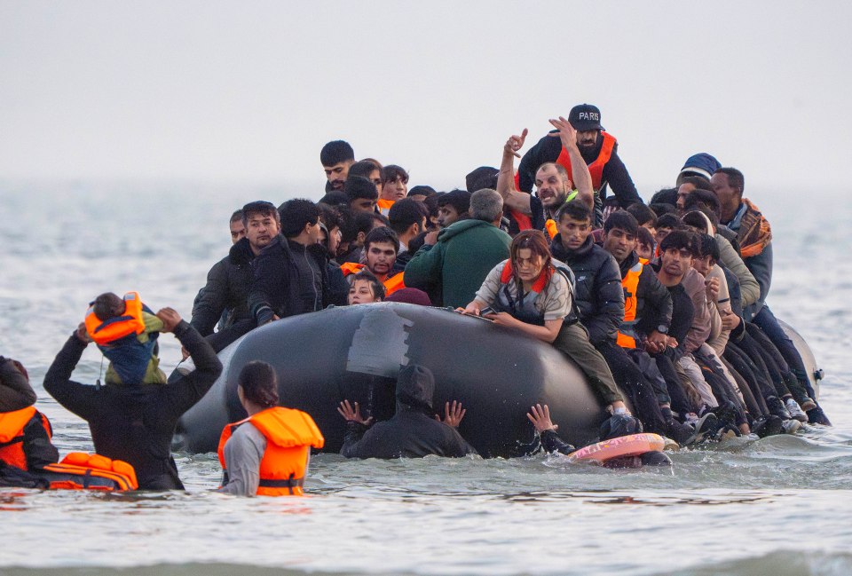 Many migrants attempting to board a small boat, some with children, are visible in the water.