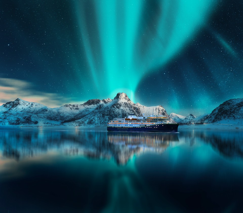 Cruise ship on water with northern lights and snow-covered mountains in the background.