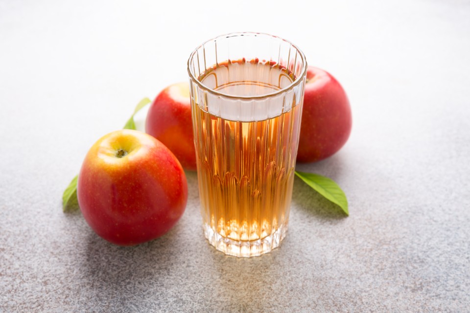 A glass of apple juice next to three red apples with green leaves on a light-colored surface.