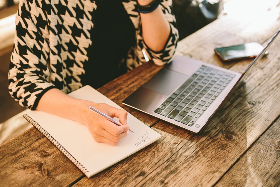 A woman writing a to-do list in a notebook on a wooden table, next to a laptop.
