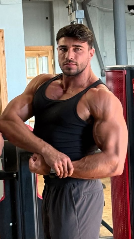 Tommy Fury posing in a black tank top and grey shorts, flexing his biceps.