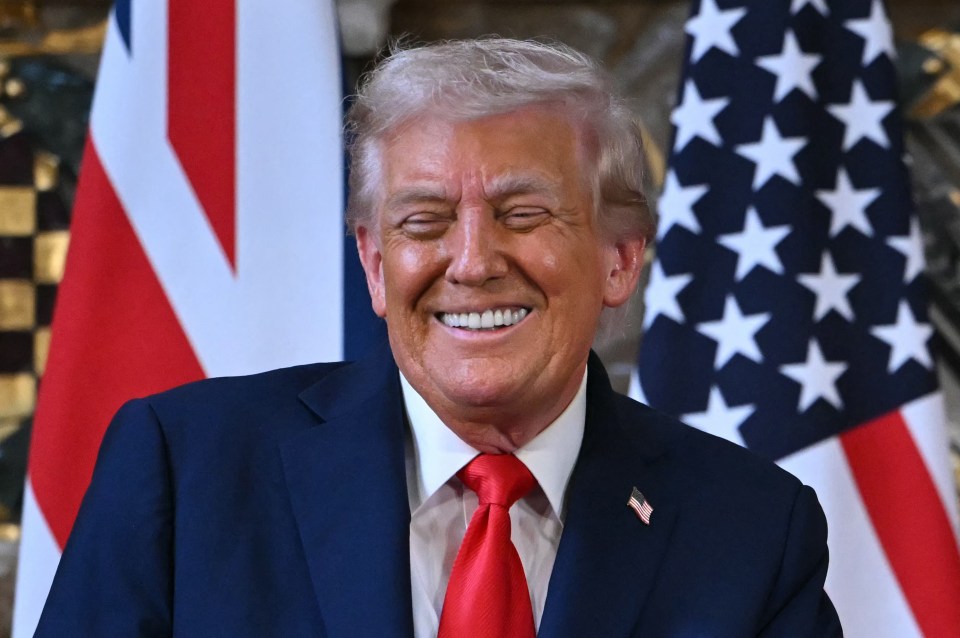 Former US President Donald Trump smiling during a press conference with flags of the UK and US in the background.