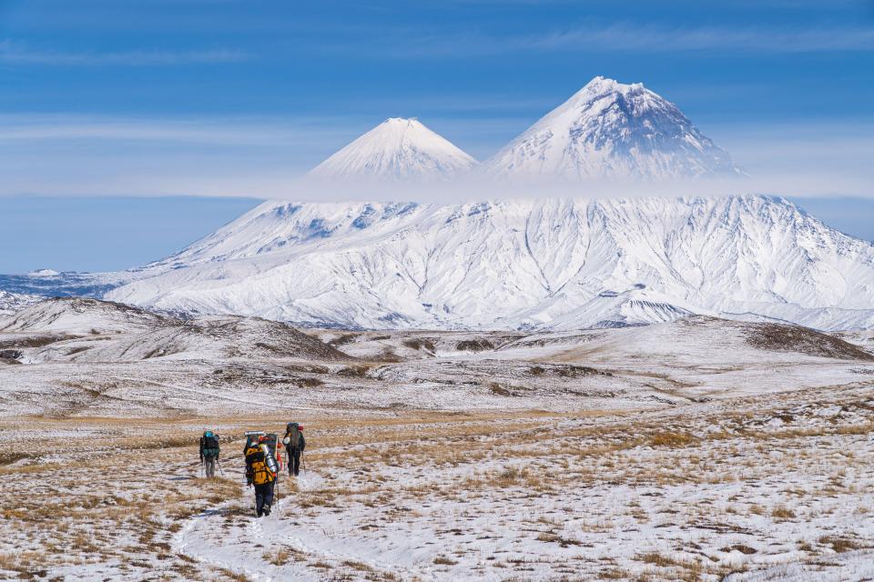 Three tourists walk across a snow-dusted, dry grass field towards the Klyuchevskoy and Kamen volcanoes in Kamchatka, Russia.