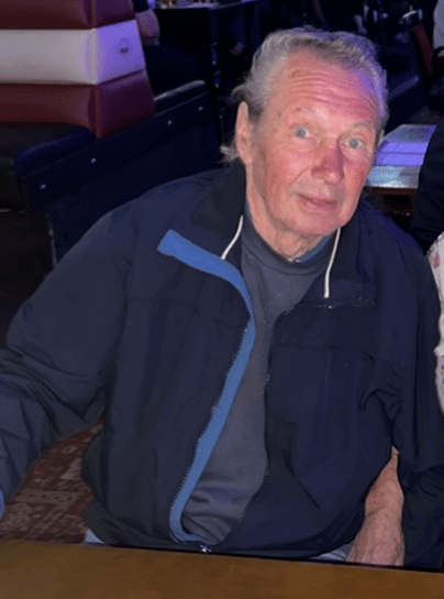A man with light eyes and grey hair wearing a dark blue jacket.