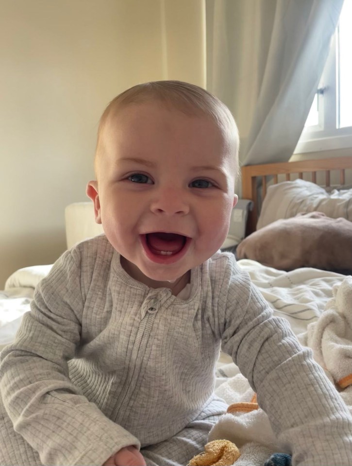 Zackary Blades, an eight-month-old baby, smiles widely while sitting on a bed.