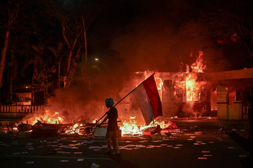 A protester with an Indonesian flag during a demonstration where a building is on fire.