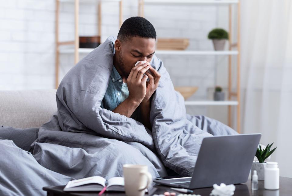 An African man wrapped in a gray blanket, sneezing into a tissue while sitting on a couch with a laptop, mug, and pills nearby.