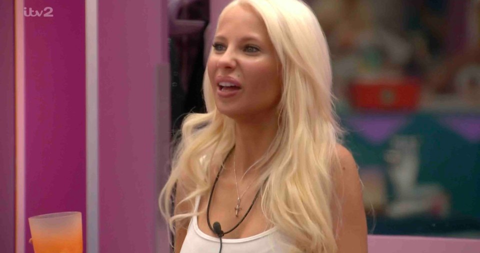 Elsa, a contestant from Big Brother, is shown in a close-up, holding an orange drink and wearing a white tank top with a microphone attached.