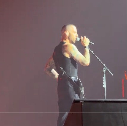 A man with a shaved head and tattoos on his arms, wearing a black tank top and pants, sings into a microphone on stage.