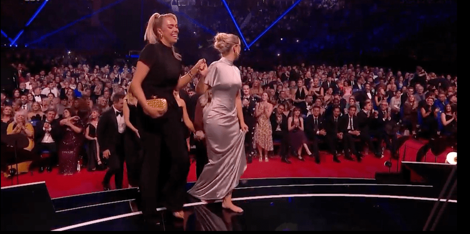 Two women walking barefoot across a stage to applause.